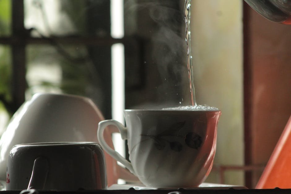 A cup of green tea with steam rising next to a bamboo matcha whisk and chawan bowl